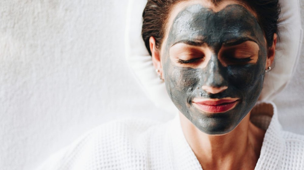 A woman rests in a robe with a charcoal mask applied to her face.