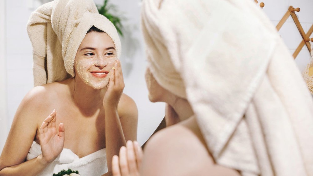 A young woman in a towel with a towel on her head washes her face with a scrub in the mirror.